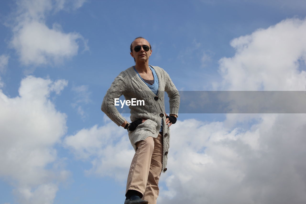 Low angle portrait of mature man with hands on hip standing against cloudy sky