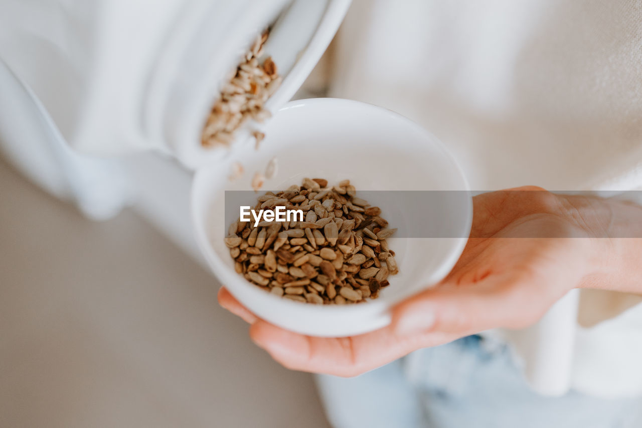 From above of crop unrecognizable female pouring sunflower seeds in bowl for healthy breakfast at home