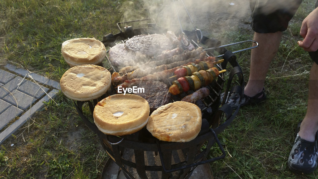 Low section of man cooking meat with vegetables and buns on barbecue grill in yard