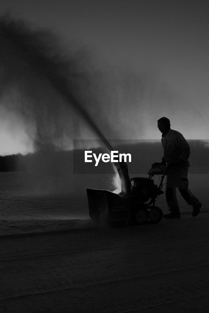 Side view of man using snowblower against sky