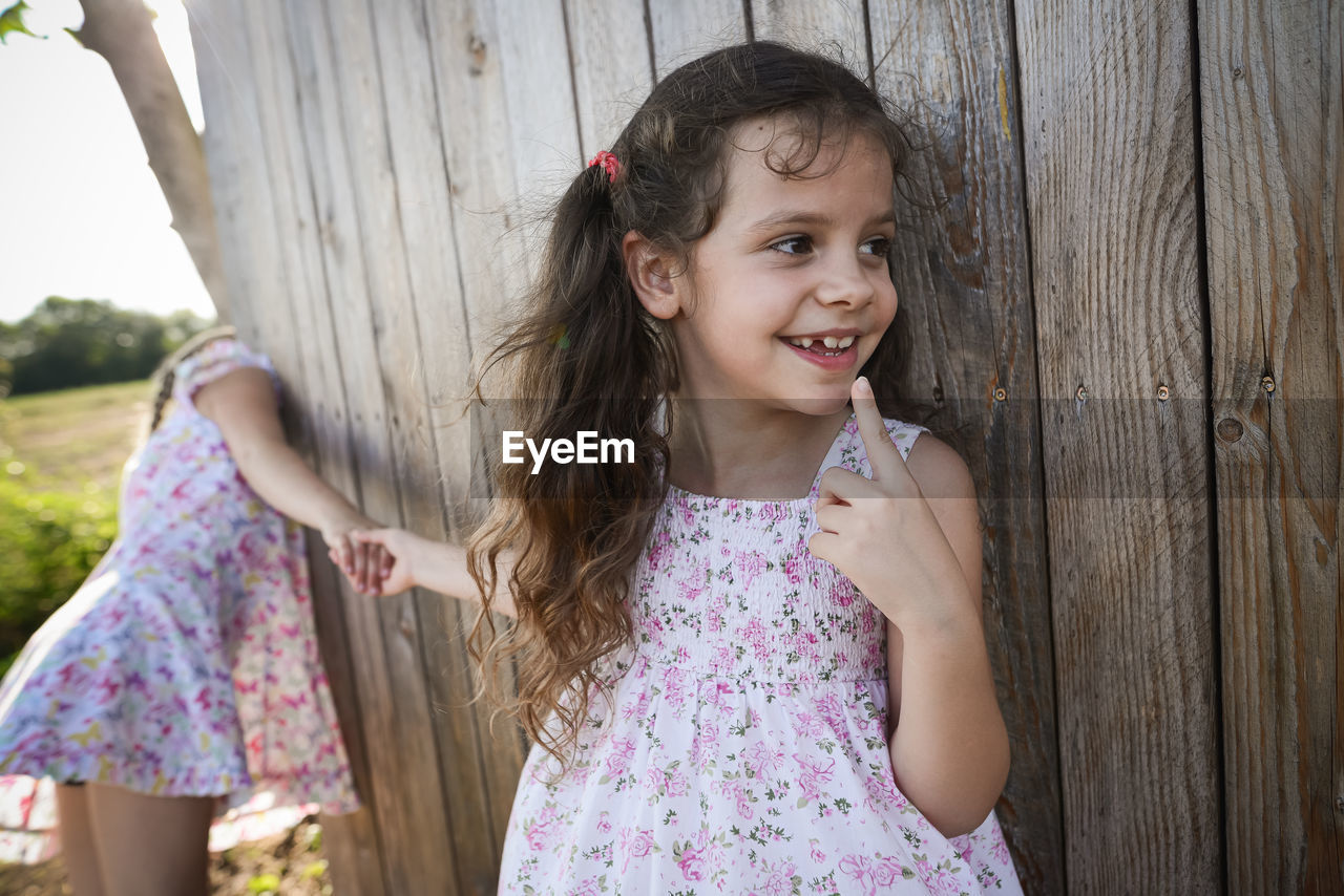 Smiling girl holding sister hands while standing against wooden fence