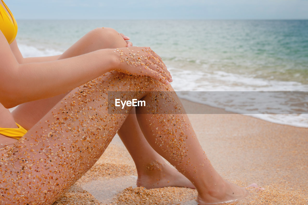 Close-up of sea waves near the feet of a young woman who slowly sits and relaxes on the beach. women