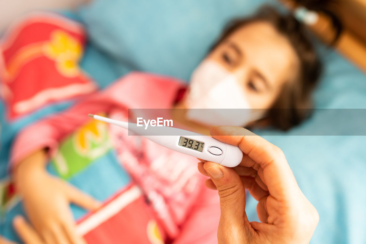 Close-up of woman hand holding thermometer over daughter lying on bed