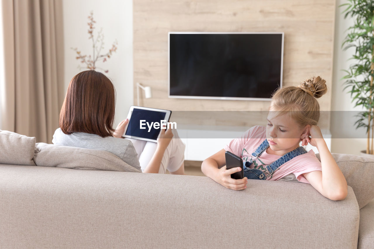 Woman holding digital tablet while sitting by daughter on sofa at home