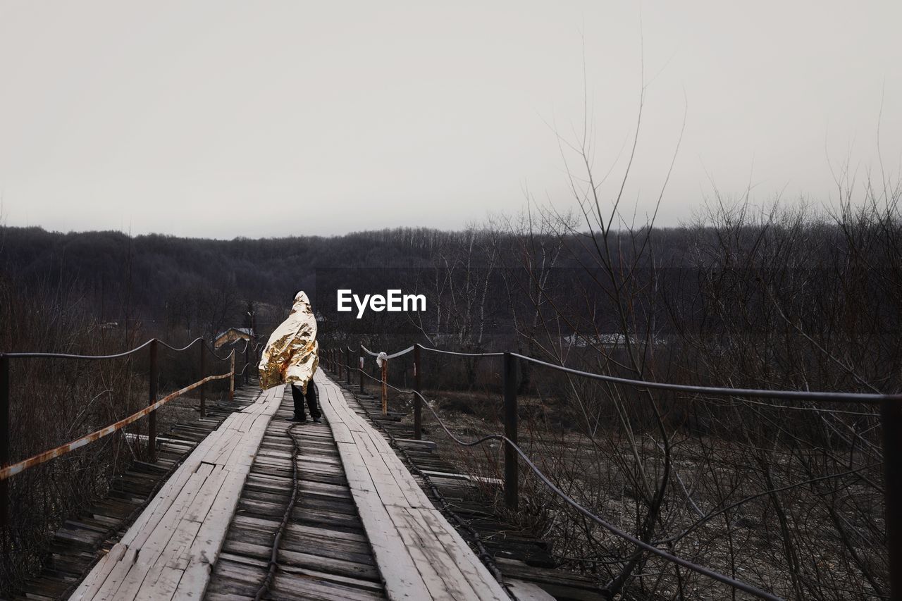 Man in raincoat standing on footbridge against sky