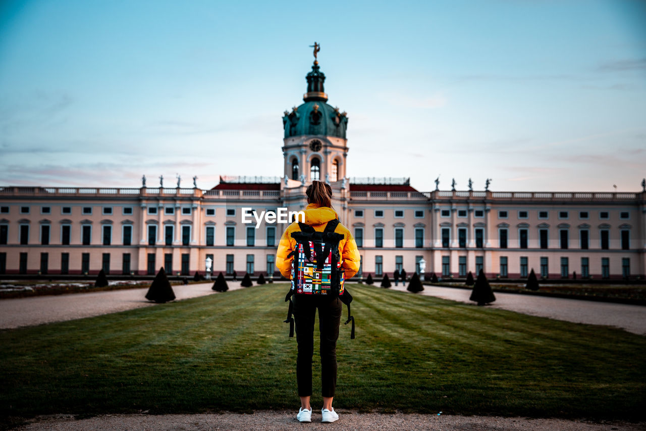 Solo female stands in front of palace with backpack with travel flag patches in evening light.