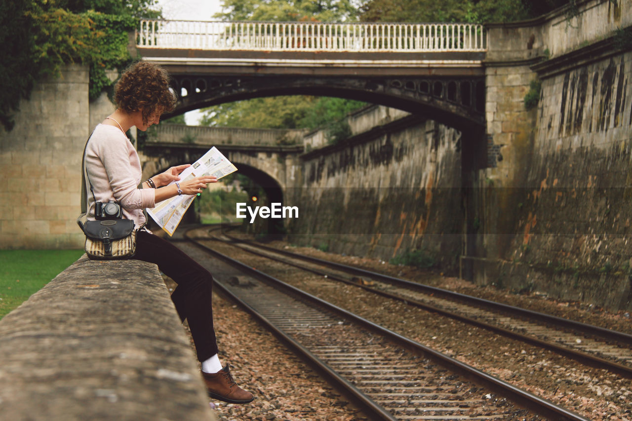 Woman sitting on retaining wall reading newspaper by railroad tracks