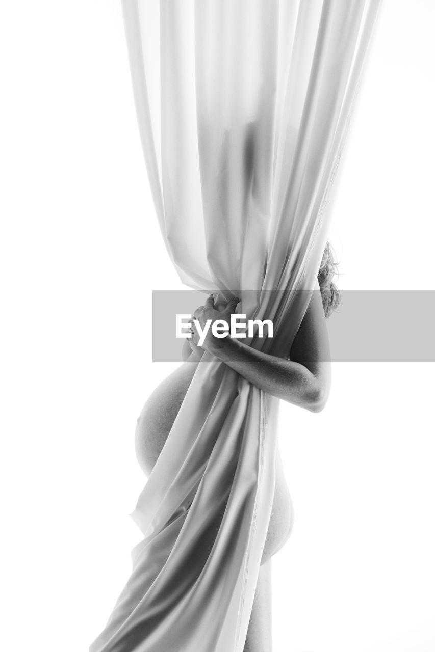 Side view of naked pregnant woman standing behind curtain against white background