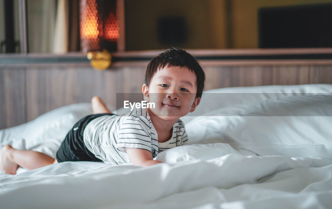 Portrait of little boy wear striped shirt feel relax, laydown on bed, looking at camera. bed vibe.