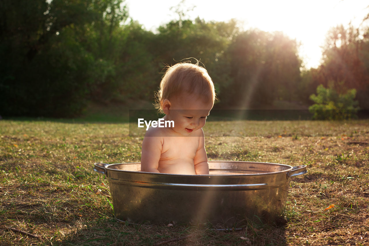 Cute little kid sits in a basin of water in nature and has rays of the setting sun, golden bathe. 