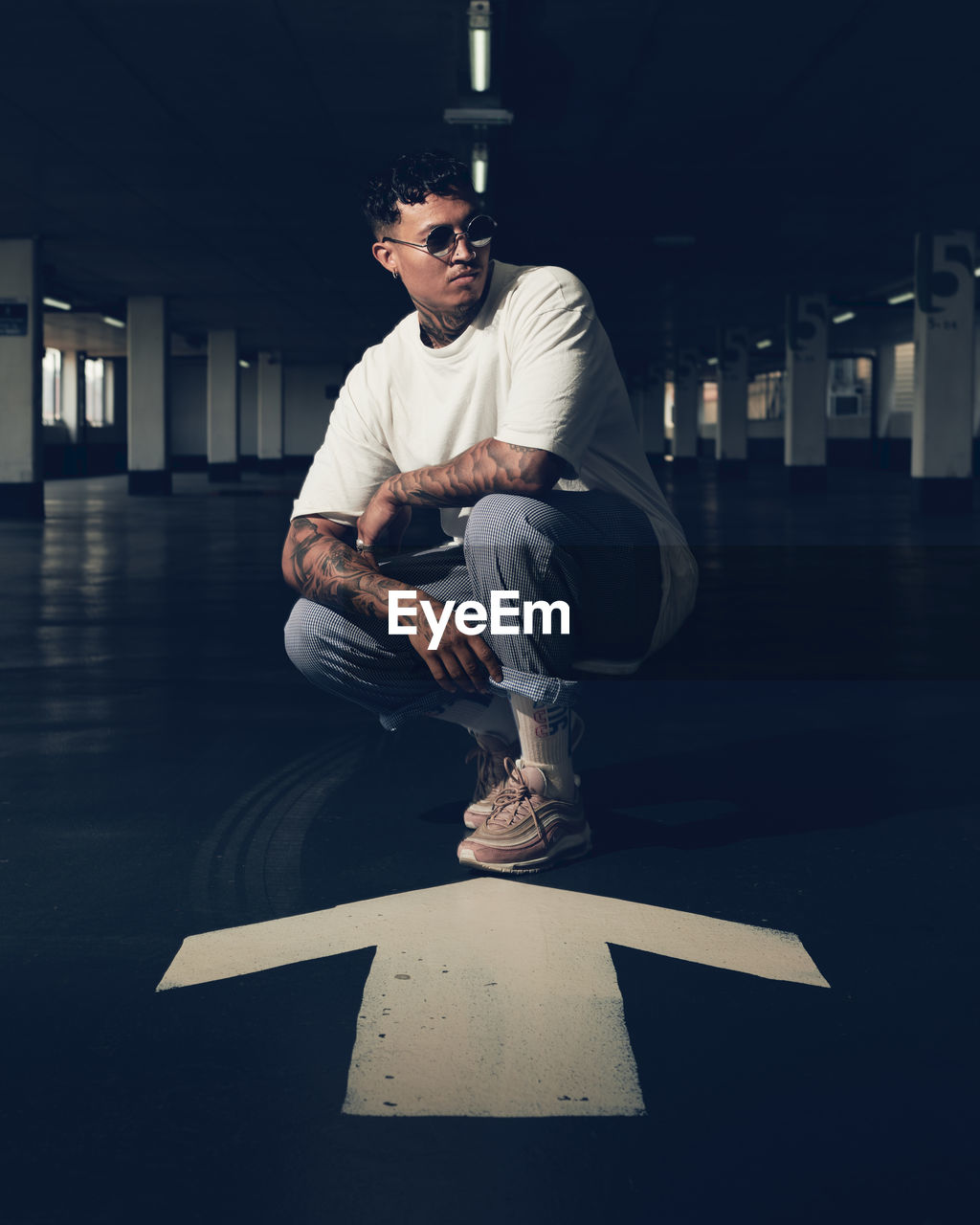 Young man wearing sunglasses crouching on arrow symbol in parking lot at night