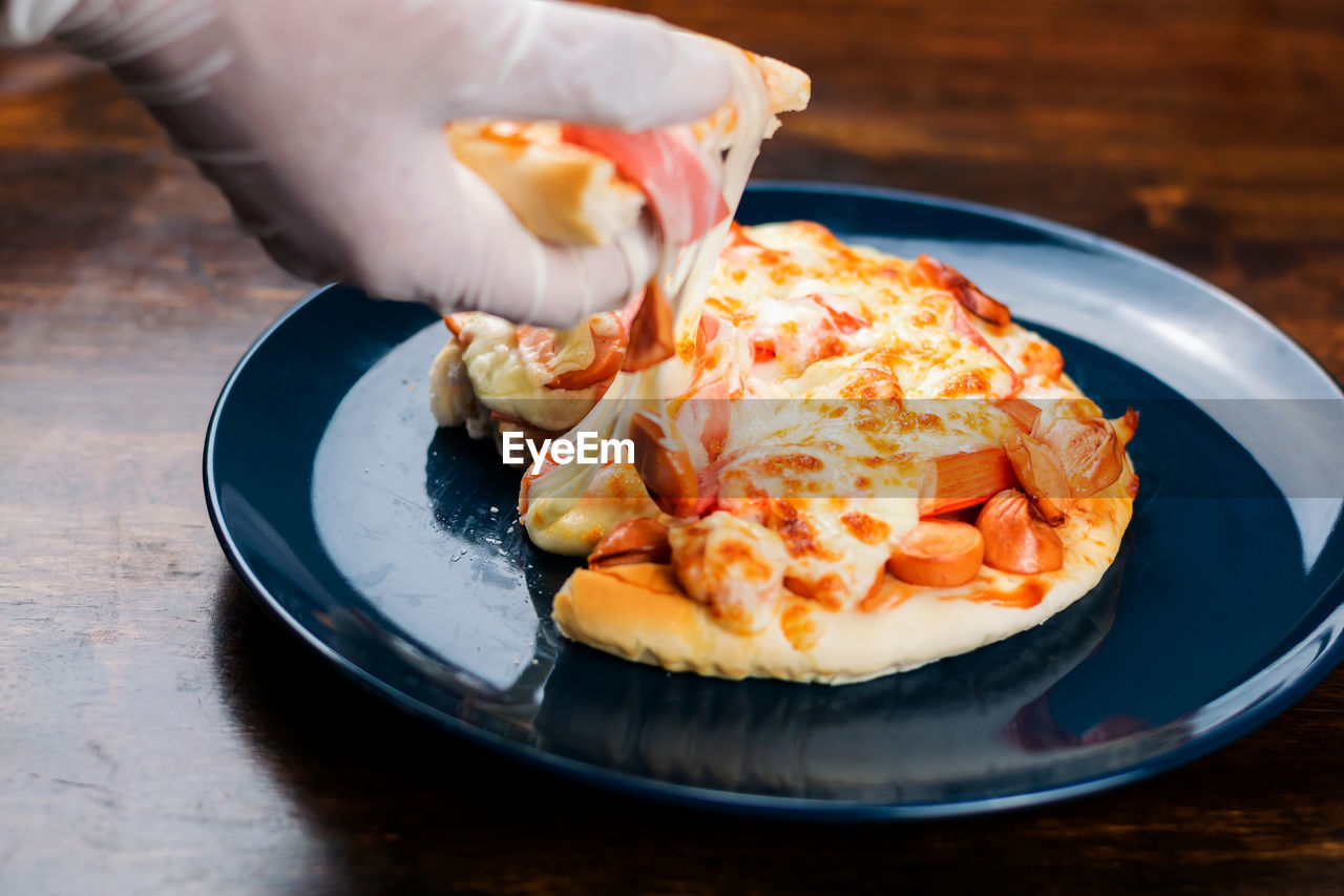 Close-up of hand wearing rubber gloves picking up pizza slice in a ceramic plate on a wooden table.