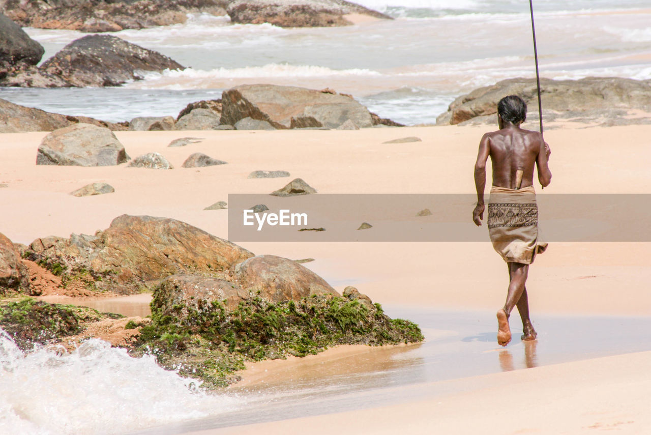 Rear view of shirtless fisherman running at beach towards sea on sunny day