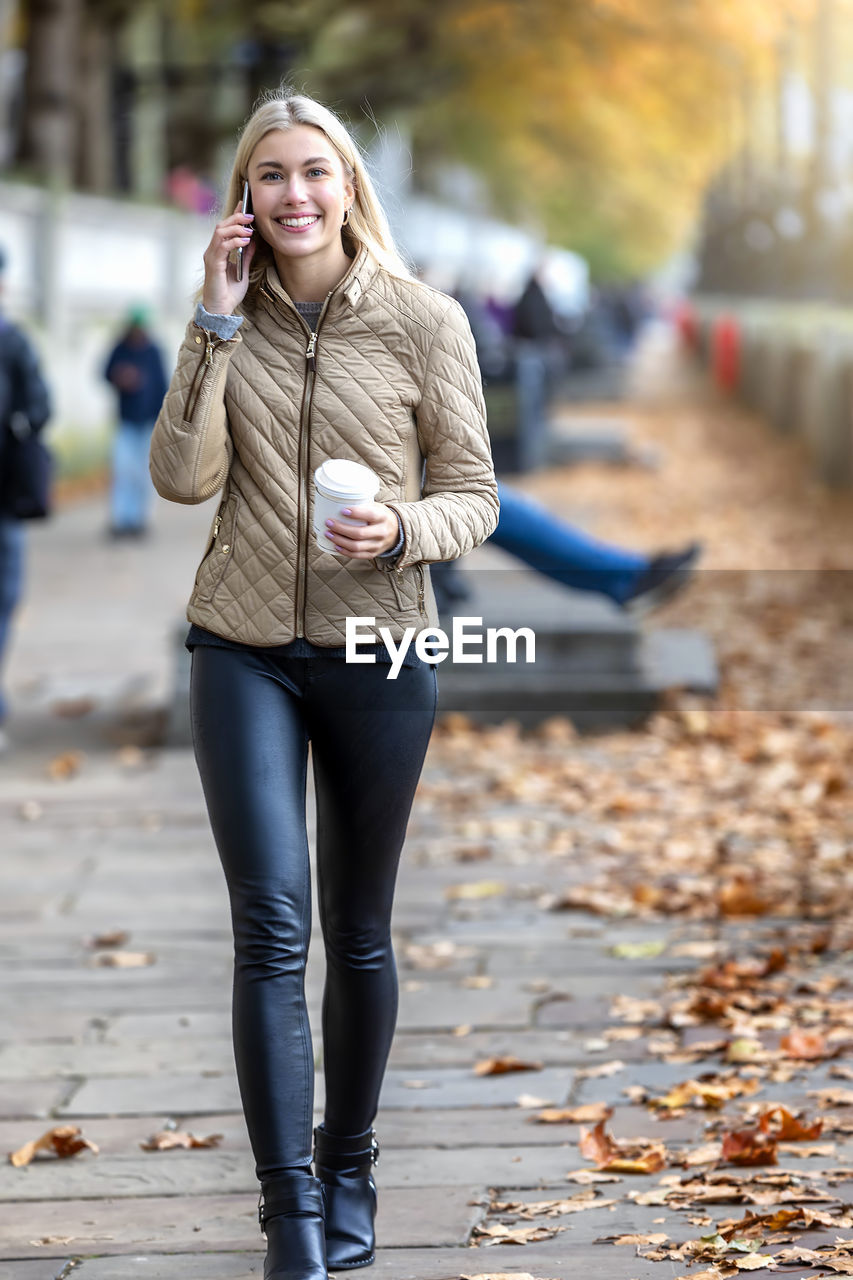 Portrait of smiling young woman talking over smart phone while walking on footpath