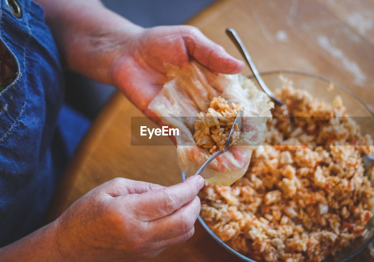 The hands of a senior woman hold a raw leaf of sauerkraut and put a metal teaspoon of rice-meat