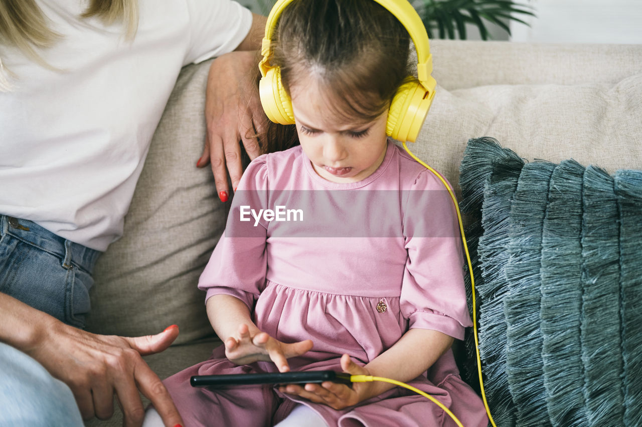 Little girl in yellow headphones sits on a sofa with a phone.