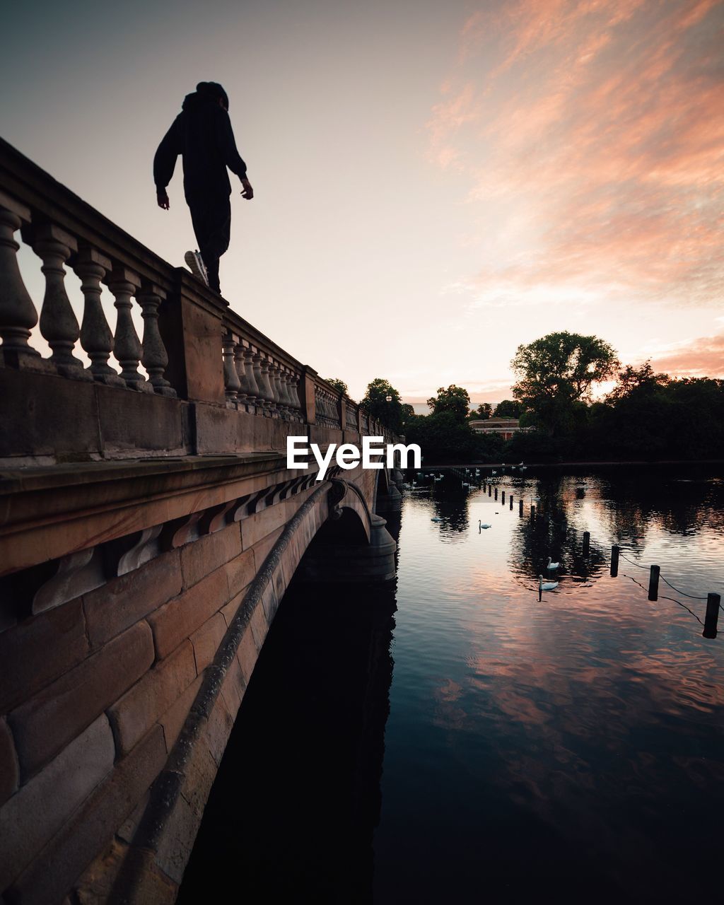 Low angle view of man walking on bridge railing over river at sunset