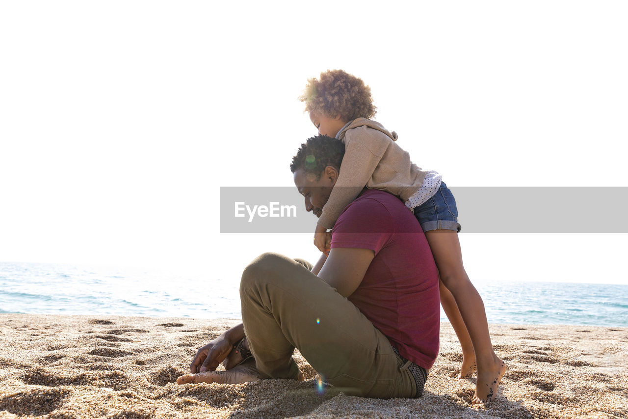 Girl embracing father sitting at beach on sunny day