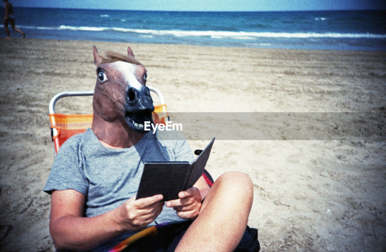 Man in horse head reading book on beach