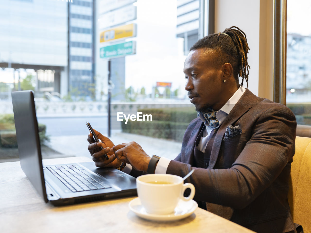 Side view of concentrated african american businessman in suit sitting at table with coffee in cafe and browsing smartphone while working on remote project