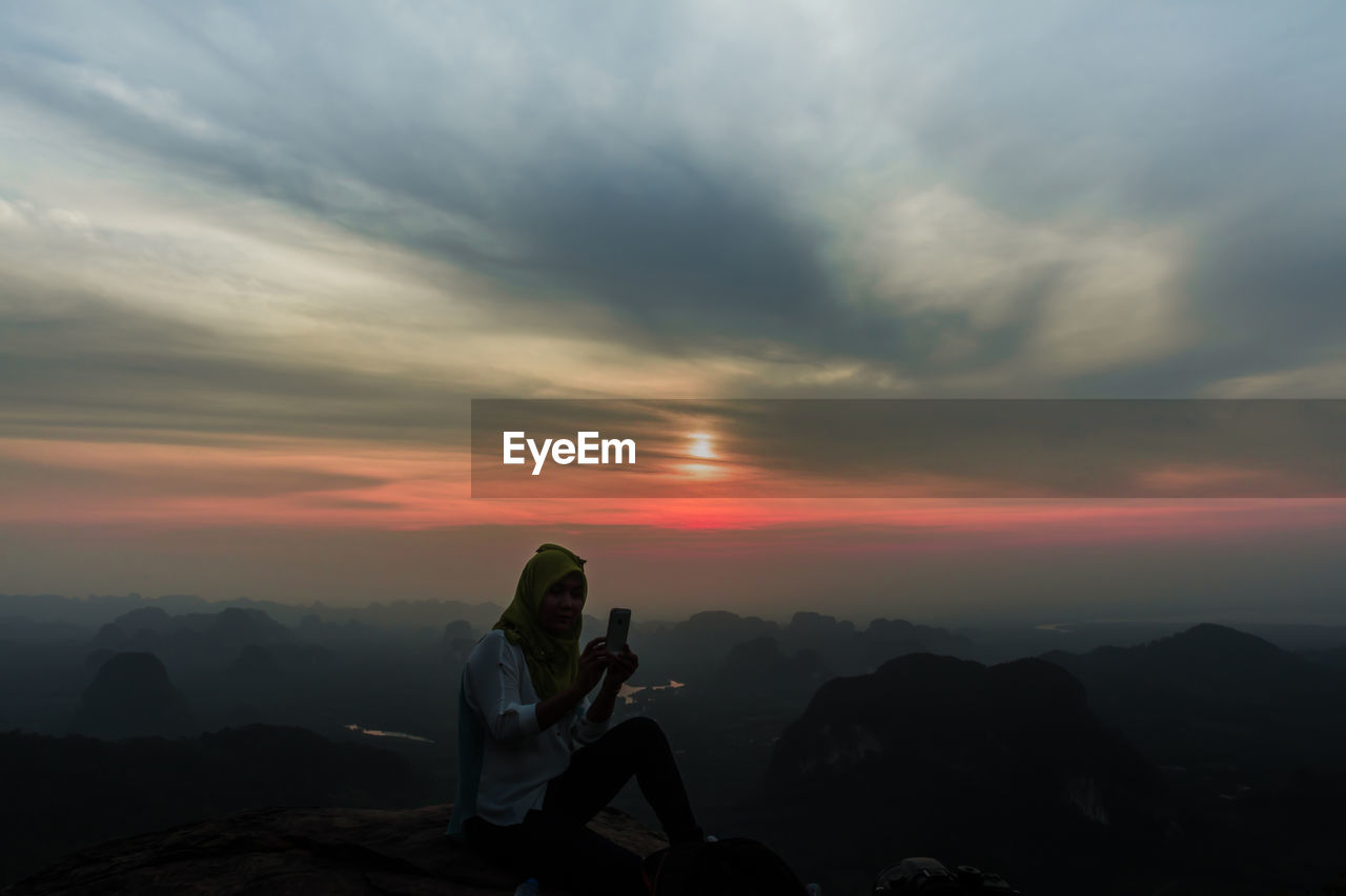 Woman photographing on mountain against sky during sunset