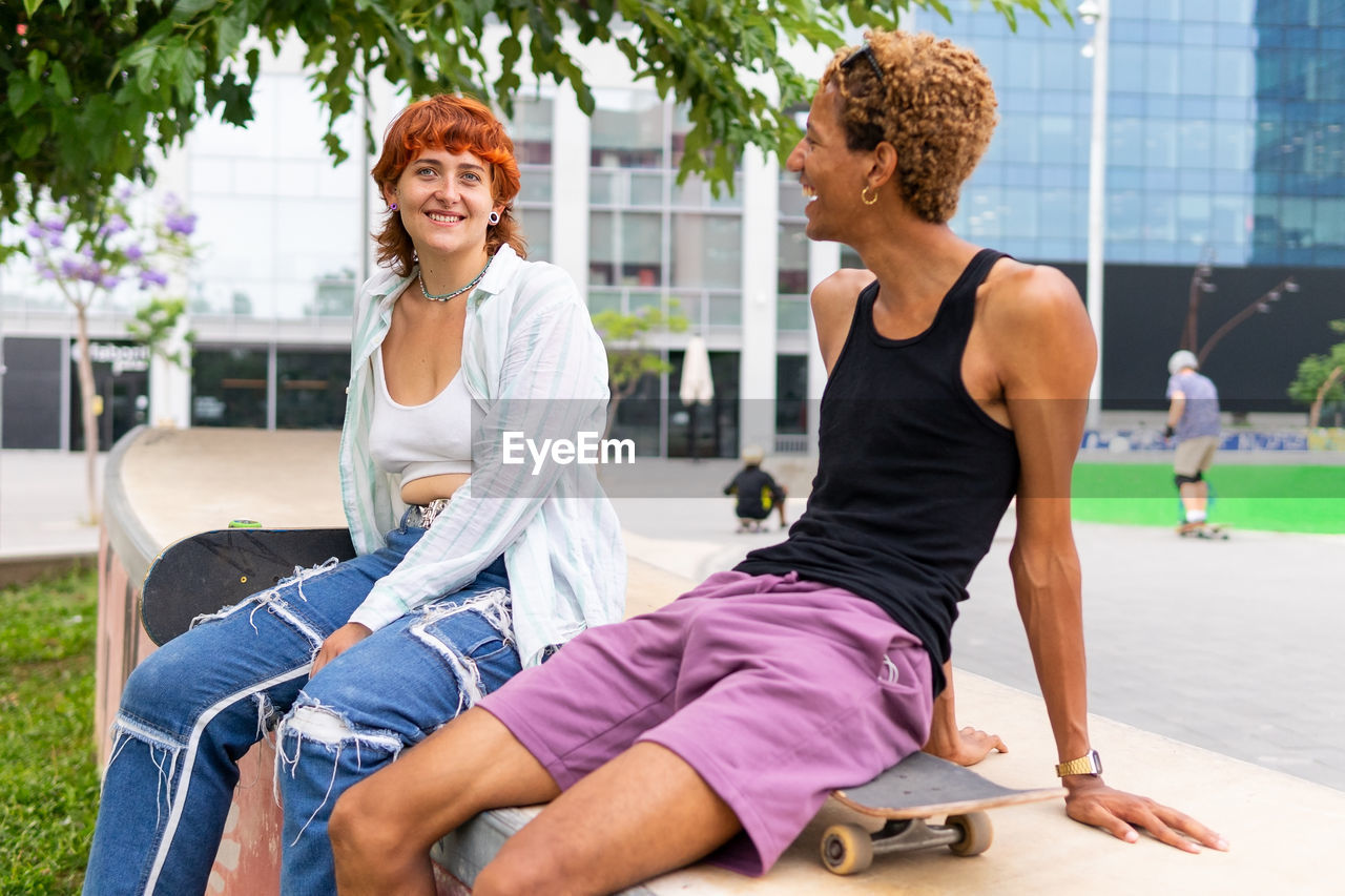 Positive multiracial friends in casual wear with skateboards sitting together on border near modern building in city on summer day