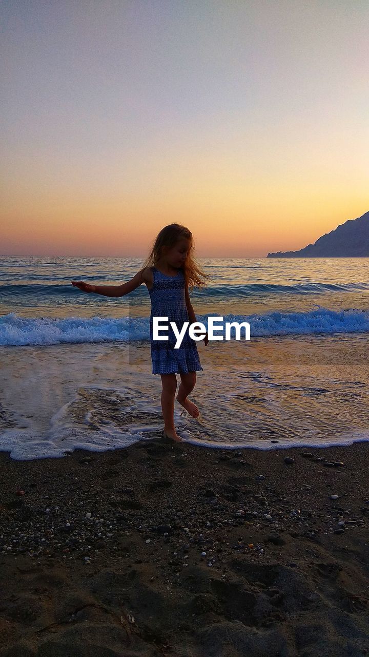 FULL LENGTH REAR VIEW OF MAN STANDING ON BEACH DURING SUNSET