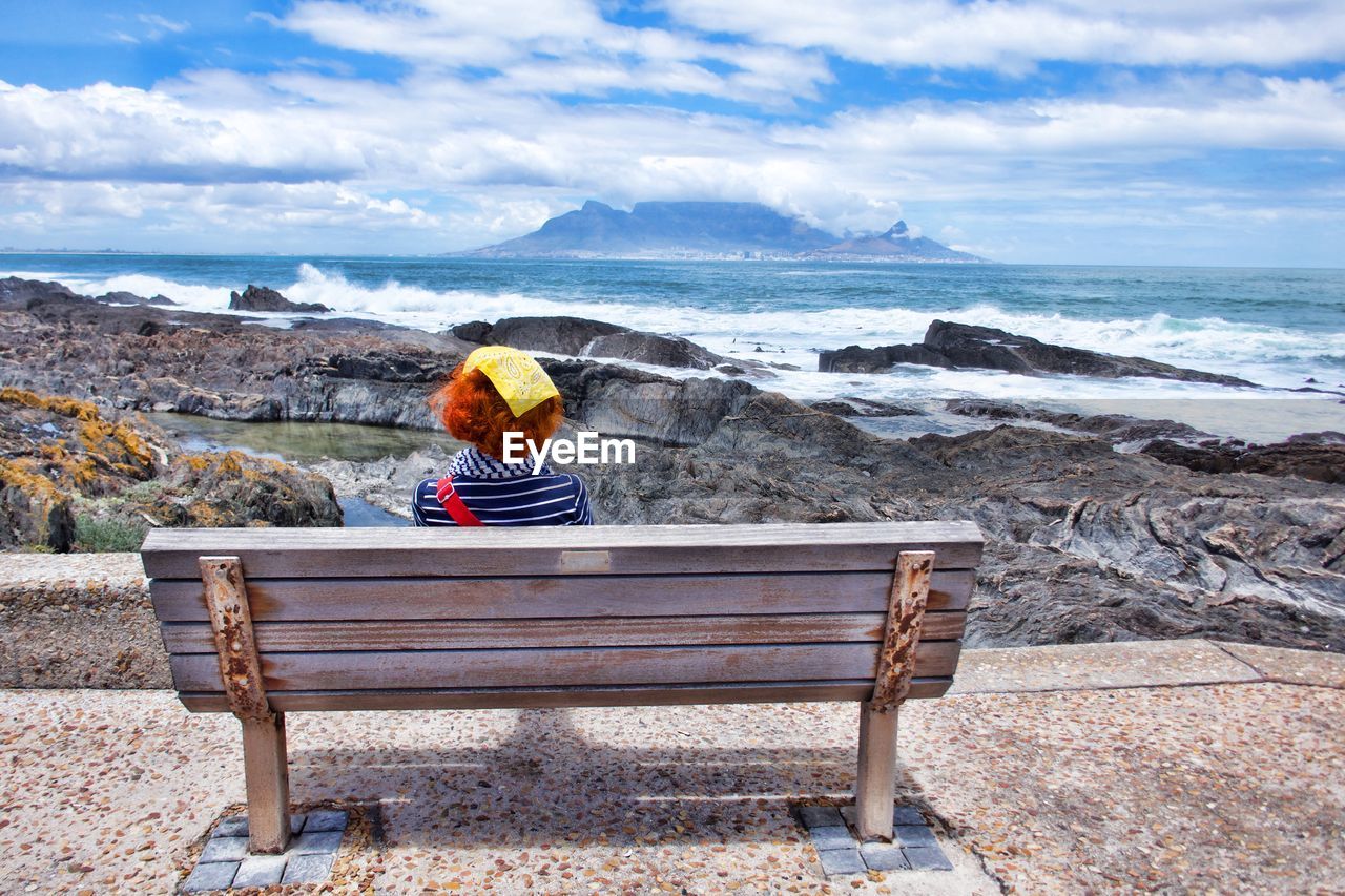 Rear view of mature woman looking at sea while sitting on bench against cloudy sky