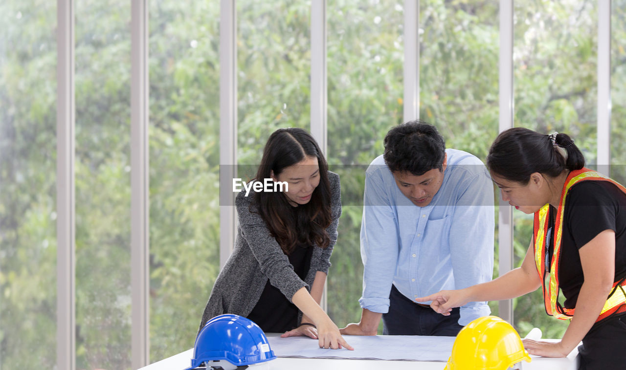 Coworkers discussing blueprint on table while standing against window in office