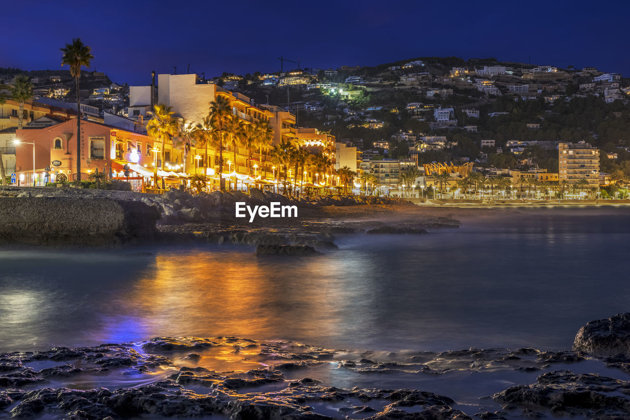 Illuminated buildings by sea against sky at night