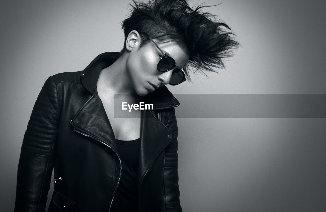 Independent serious youthful woman with short dark hair and sunglasses wearing biker jacket and looking down in studio on gray background