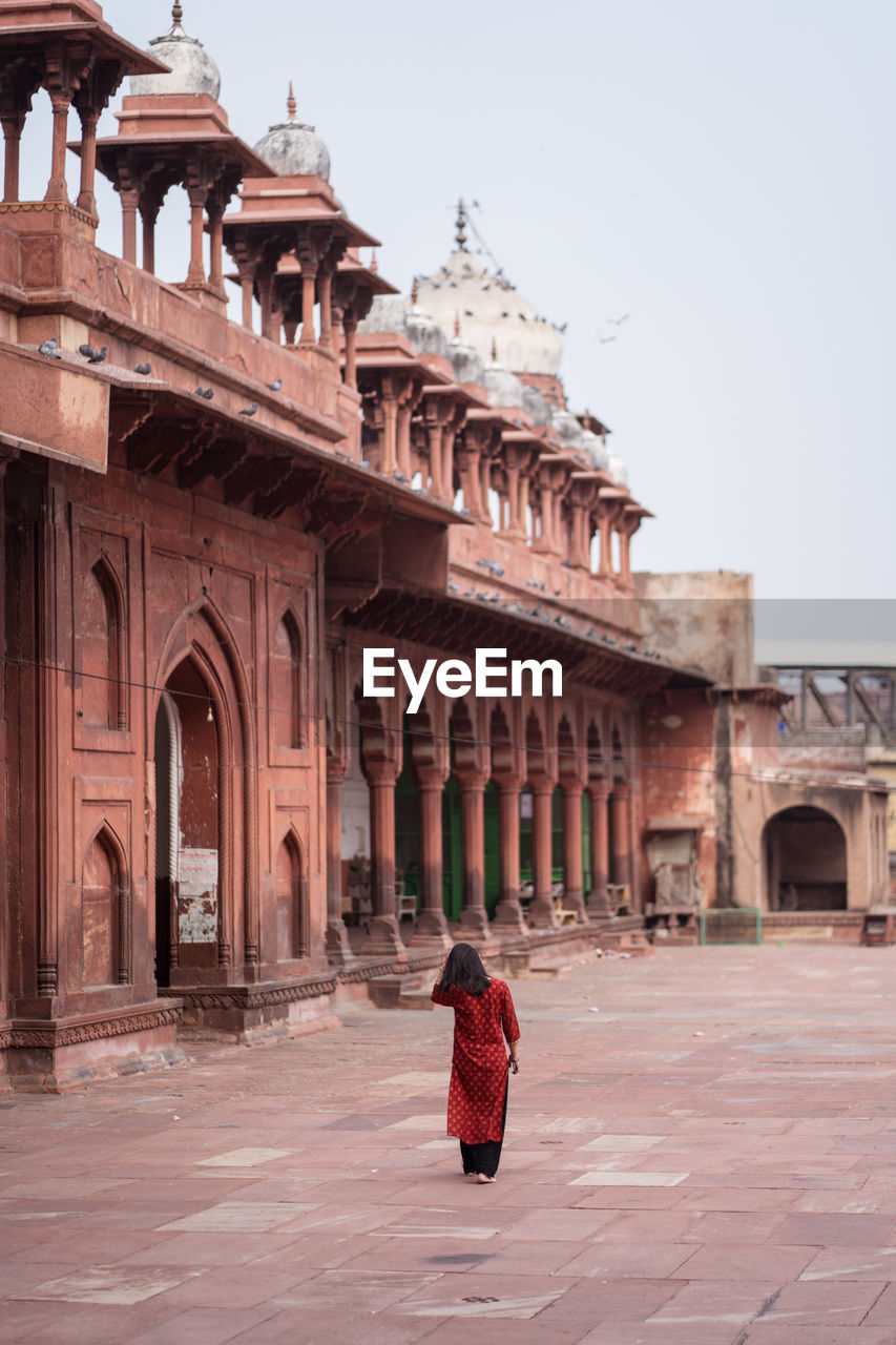 Back view of unrecognizable woman walking barefoot in weathered courtyard of ancient jama masjid building with ornamental arches while visiting agra in uttar pradesh, india