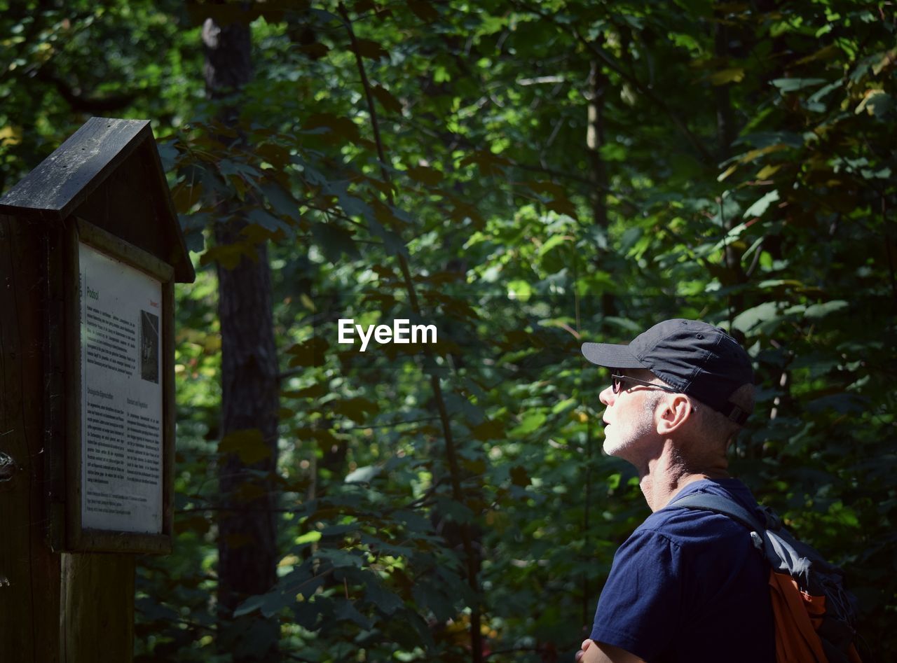 Side view of man wearing cap reading information sign in forest