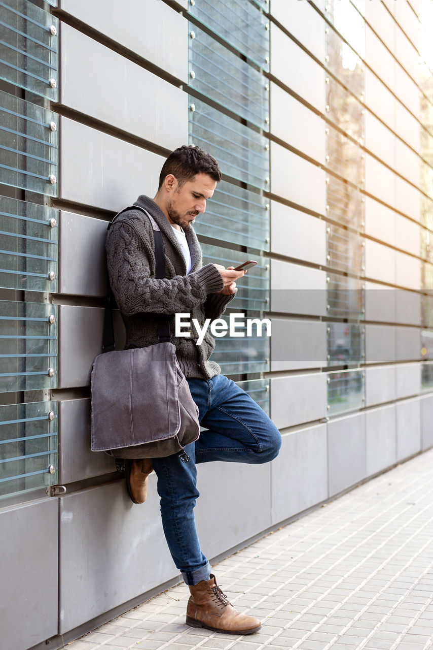 Side view of a bearded man using phone leaning on office building wall