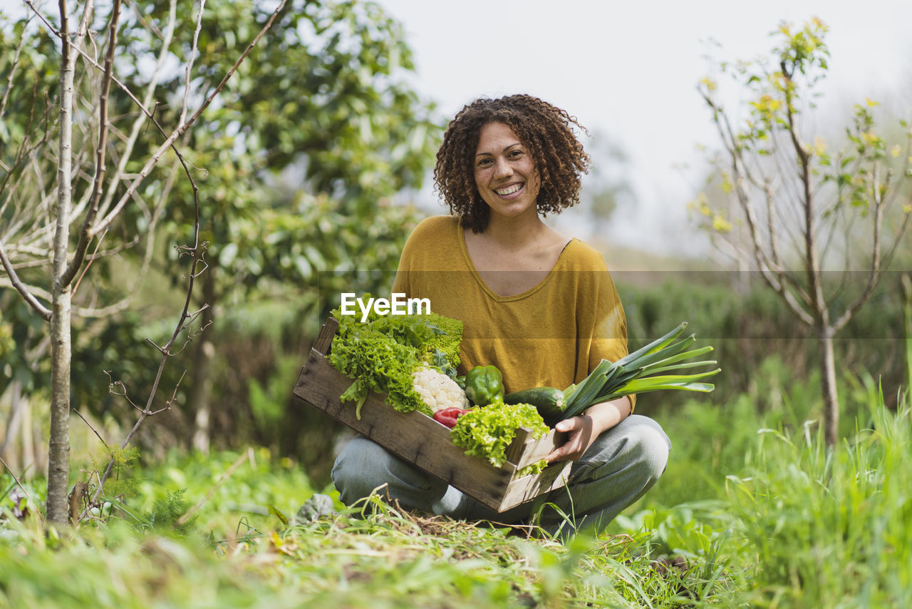 Smiling curly haired woman holding wooden crate of vegetables while squatting in garden
