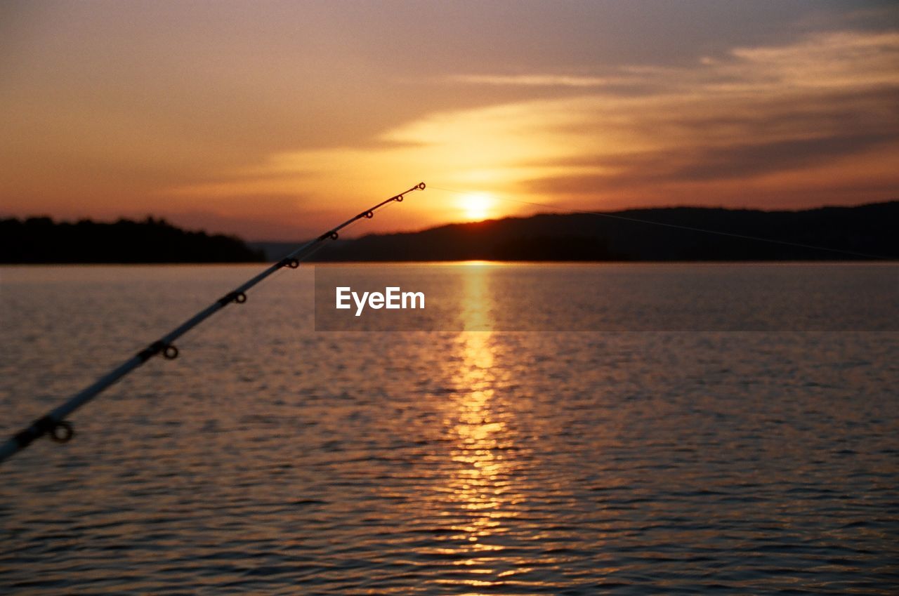 Close-up of fishing rod over lake against sky during sunset