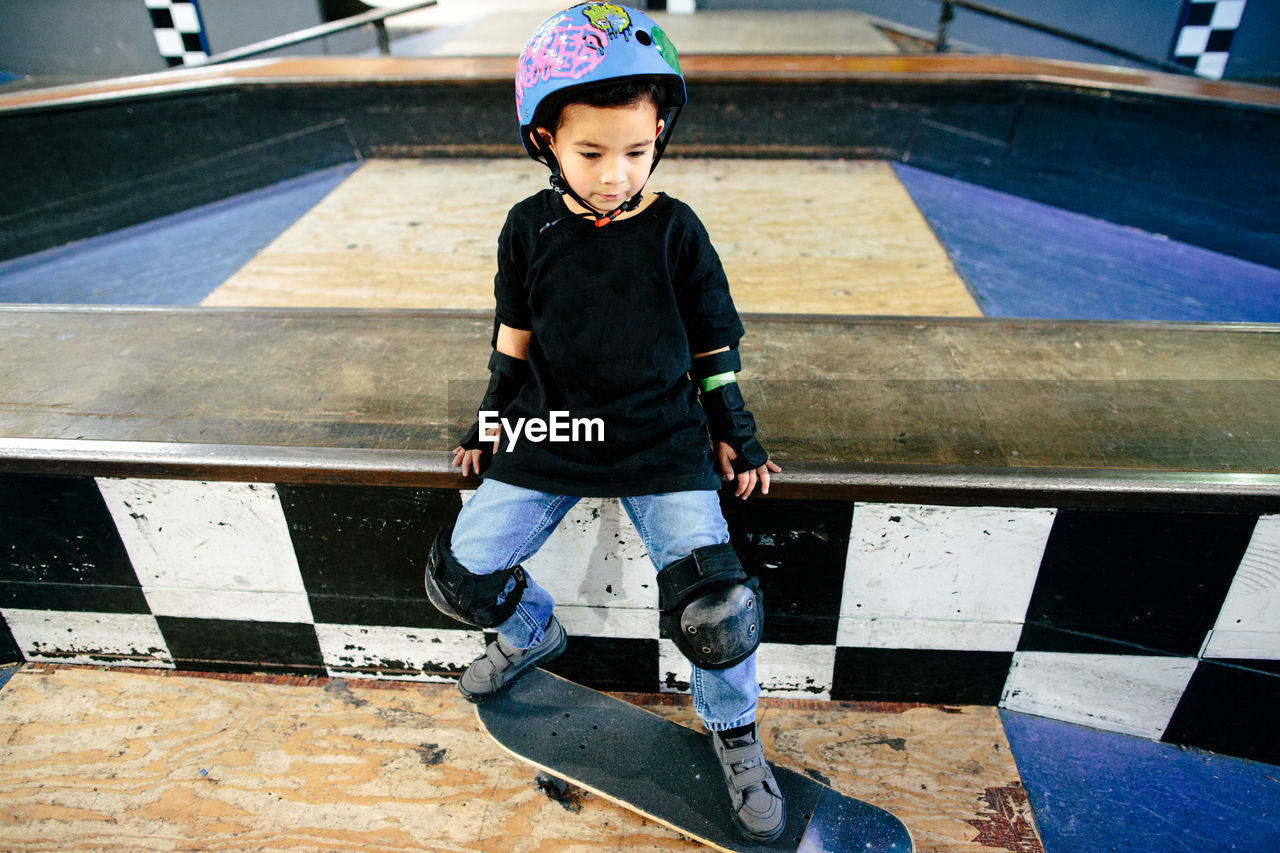 Young boy sits with his skateboard in an indoor skatepark