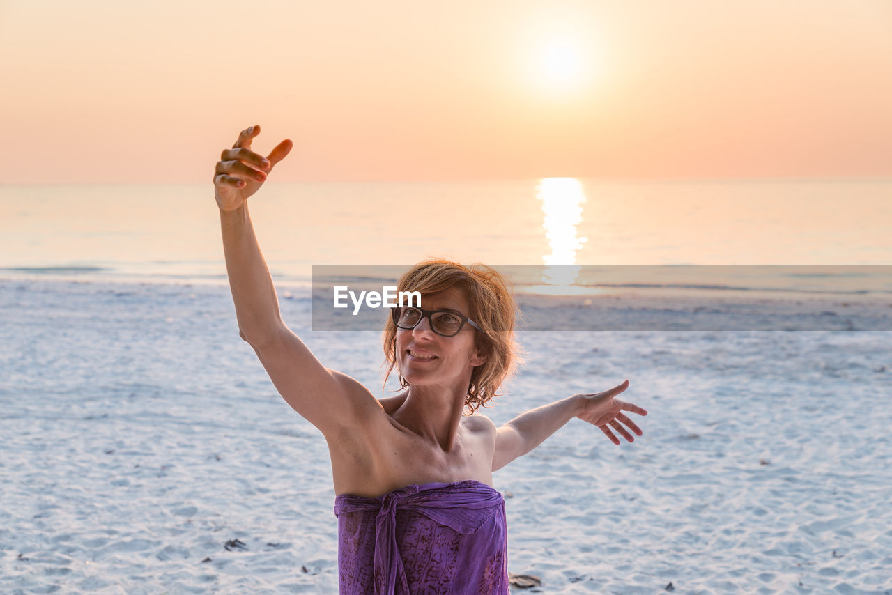 Woman with arms outstretched at beach against sky during sunset