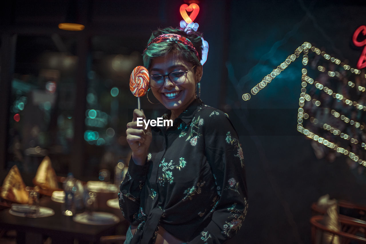 Smiling woman holding lollipop while standing in illuminated city at night