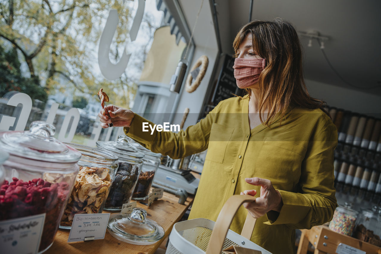 Mature female customer in mask picking up food while shopping in zero waste store during pandemic