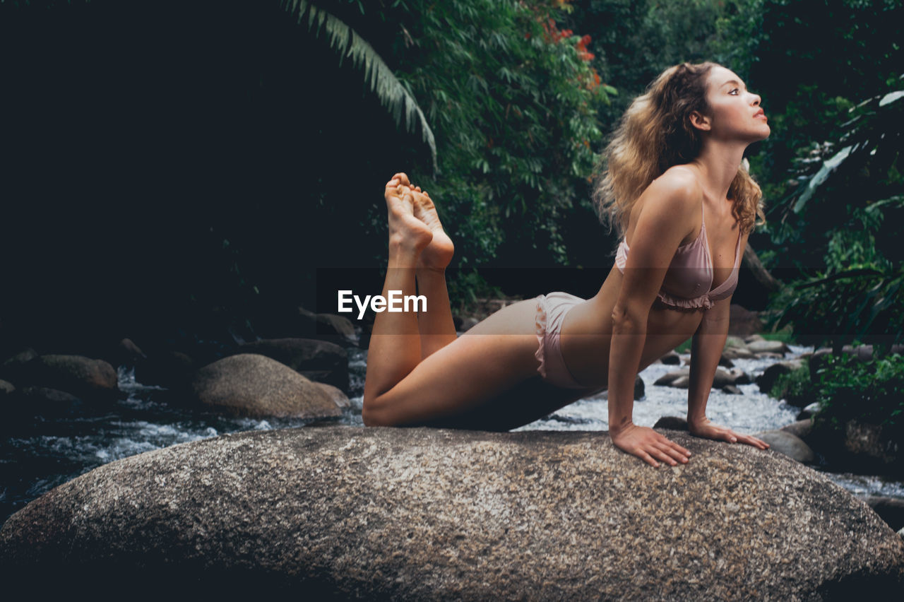 Full length of young woman wearing bikini while exercising on rock at forest