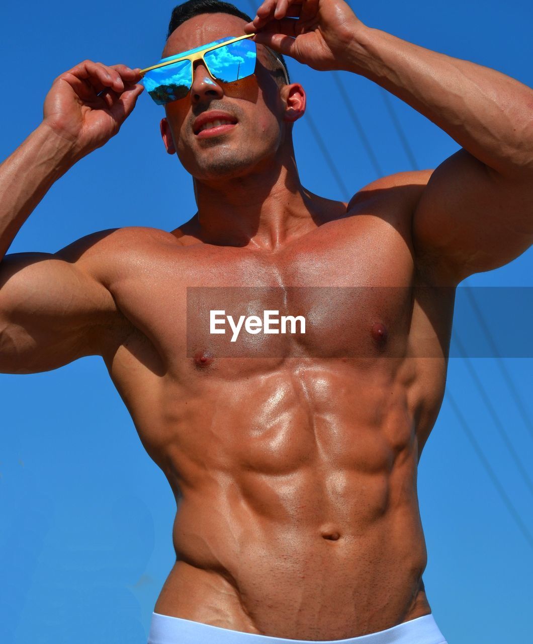 Low angle view of shirtless young man wearing sunglasses while standing against clear blue sky