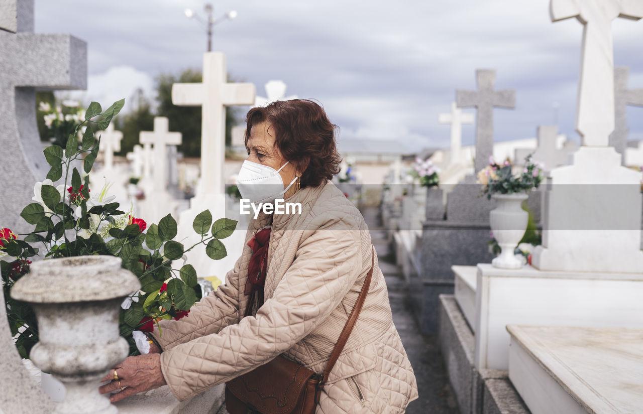 Senior woman wearing protective face mask putting flowers on grave in cemetery