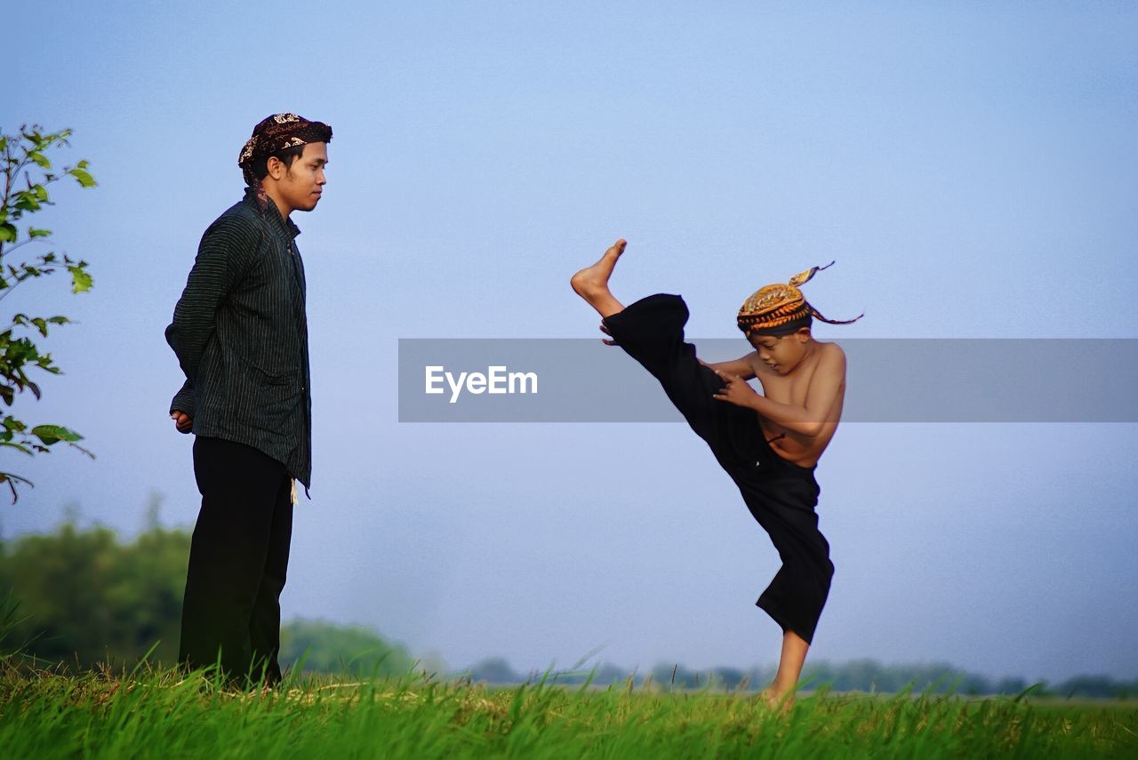 Male instructor watching at boy practicing kung fu on field against sky