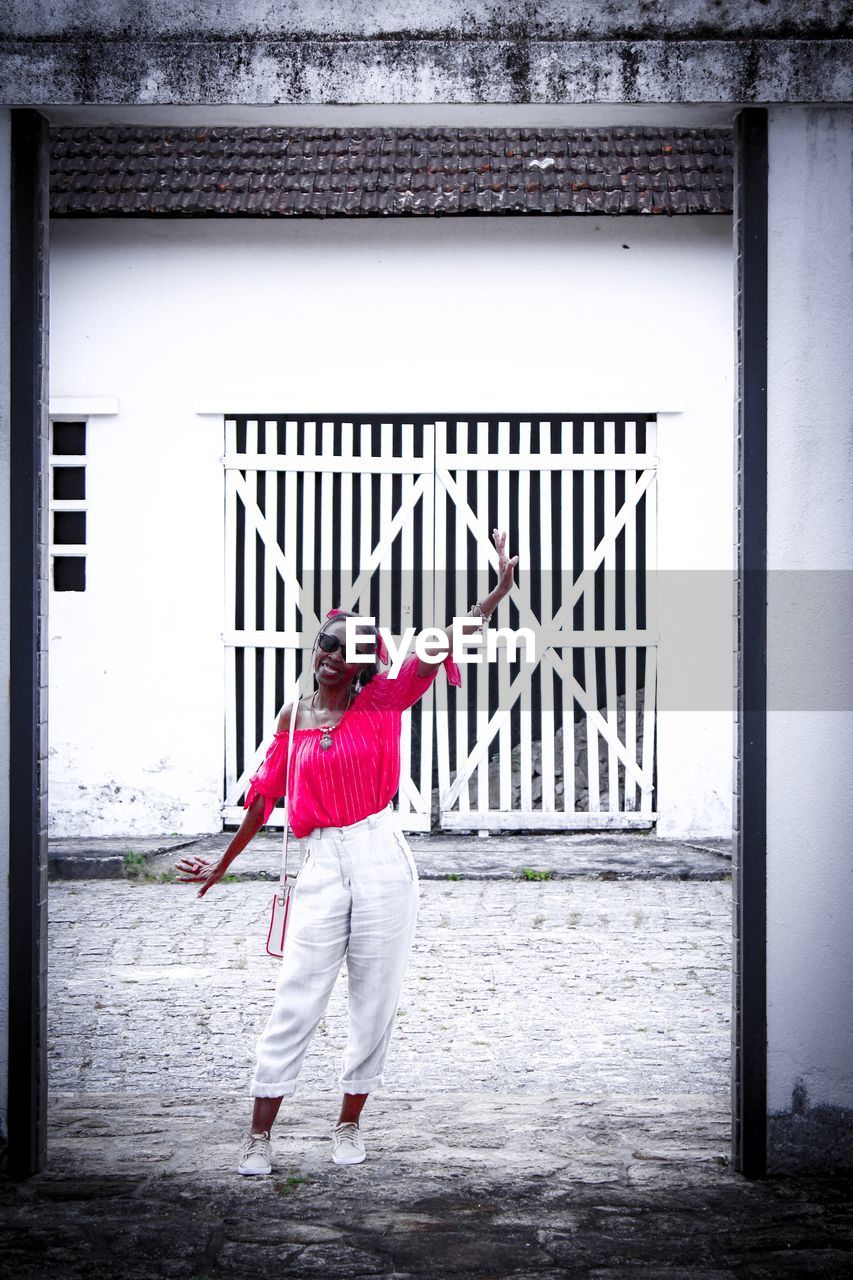 Portrait of smiling woman with hand raised standing on road against building