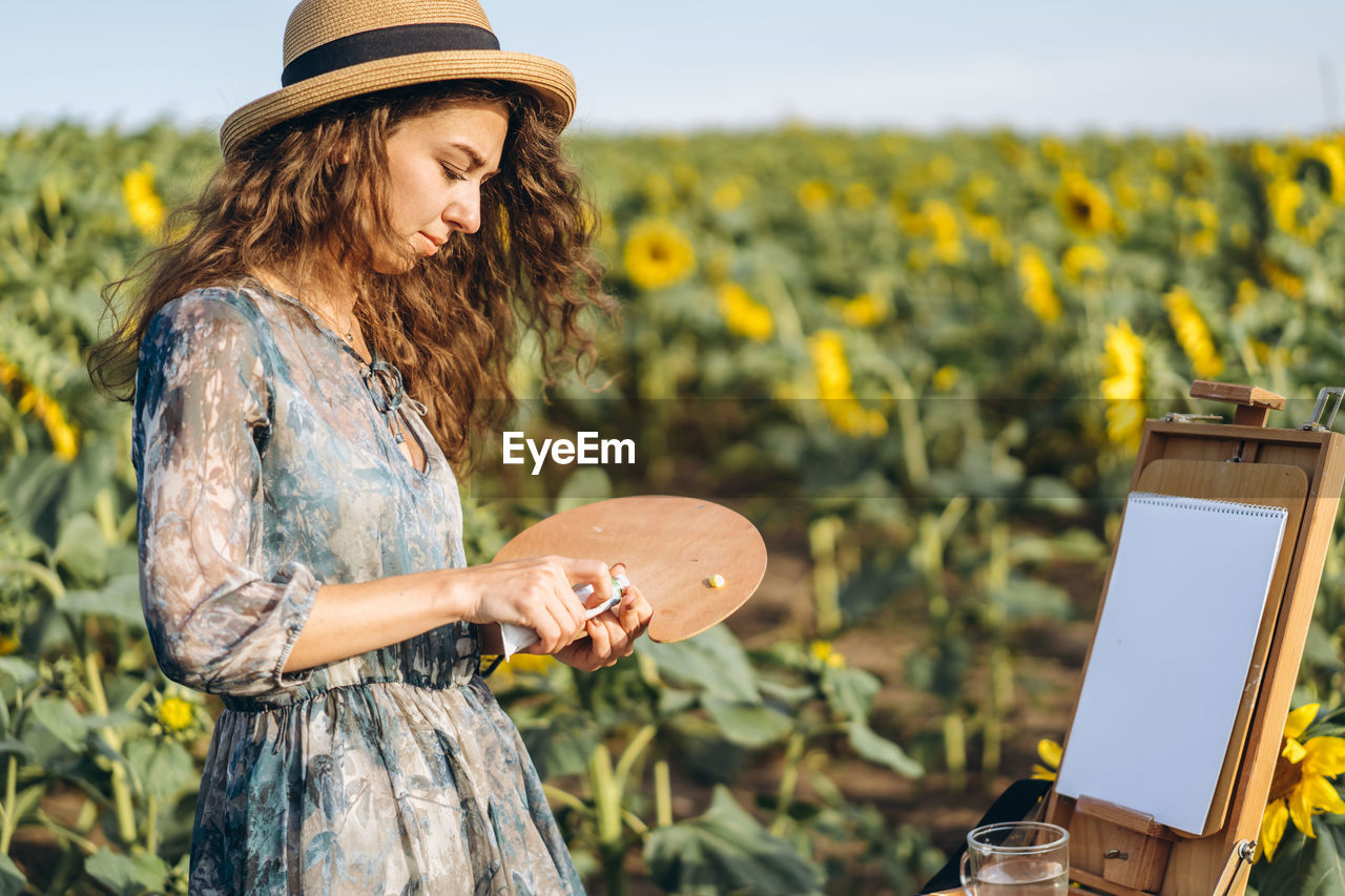 A young woman with curly hair and wearing a hat is painting in nature. 