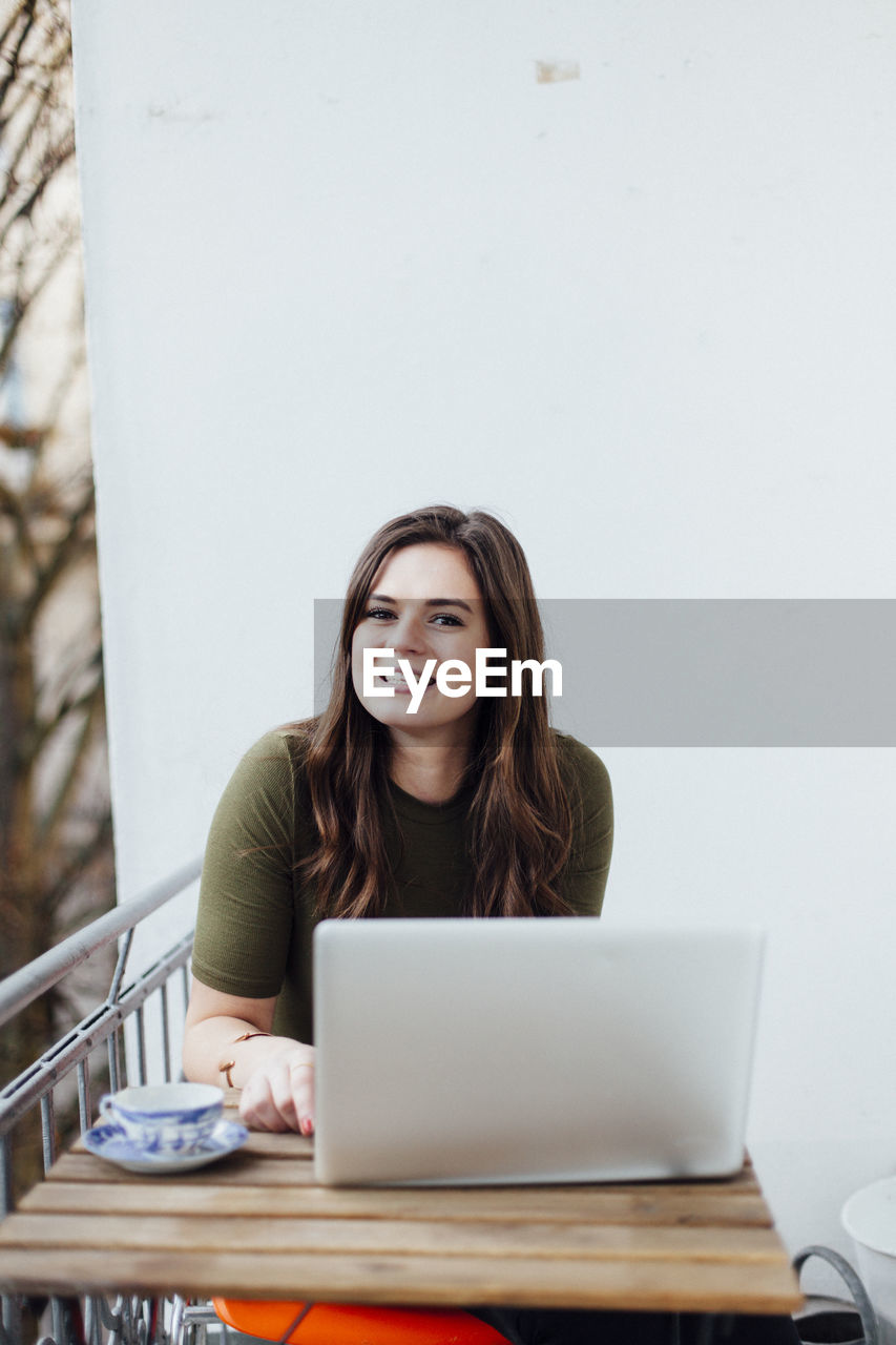 Portrait of smiling beautiful woman sitting by laptop in balcony