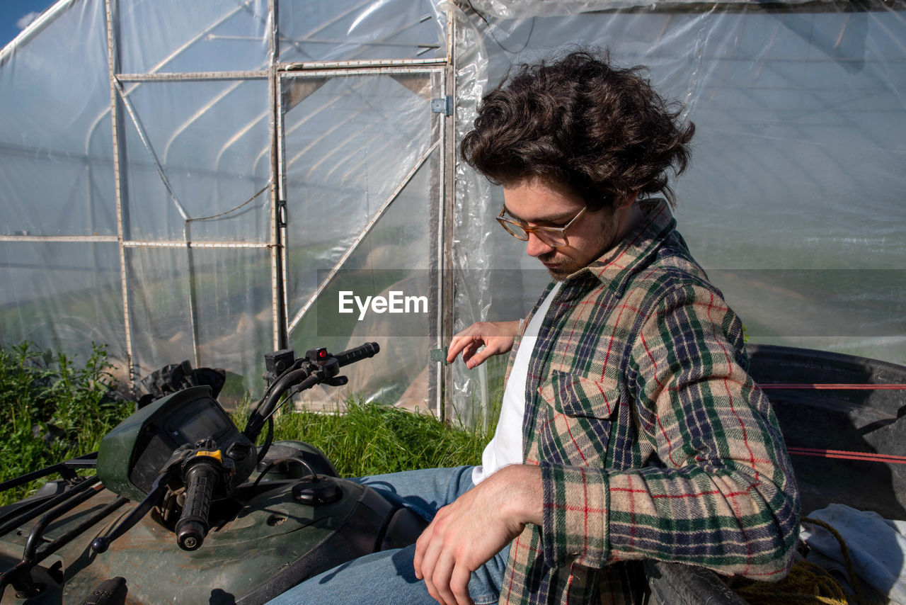 Young farmer in flannel shirt contemplates on stopped atv outside vegetable greenhouse