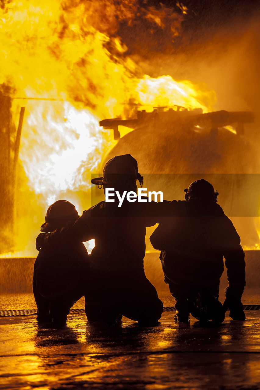 Rear view of silhouette firefighters crouching against fire at night