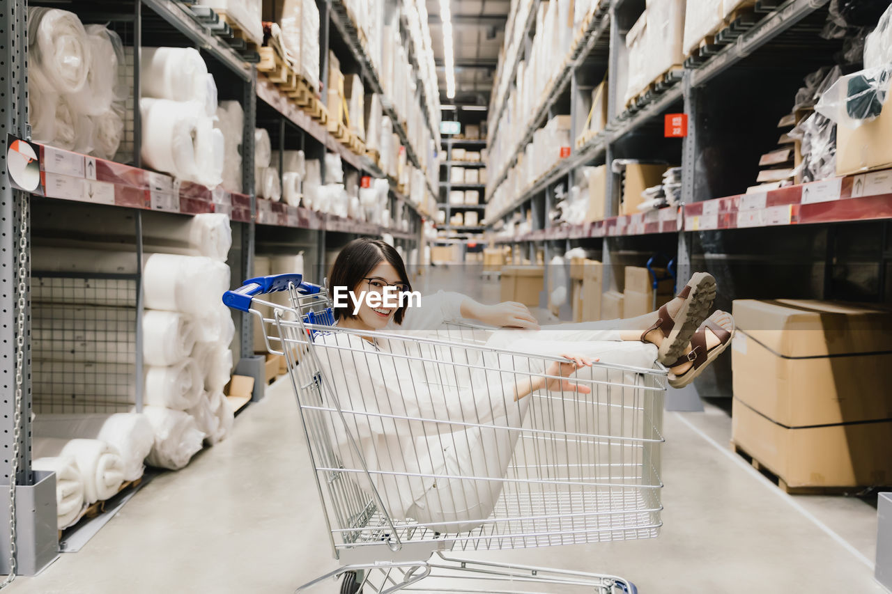 Asian young woman wearing glasses sitting in shopping cart at warehouse furniture shopping store